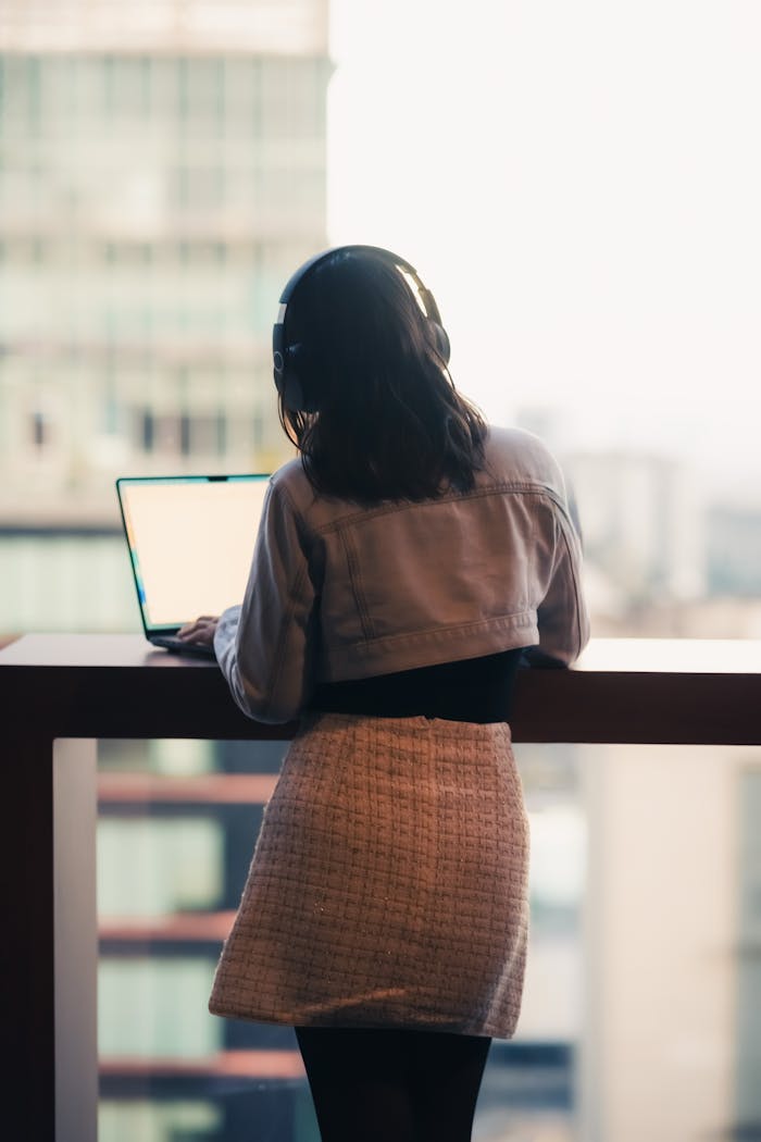 gallery-6 Young woman with headphones using laptop on a balcony in an urban setting. Perfect for remote work themes.