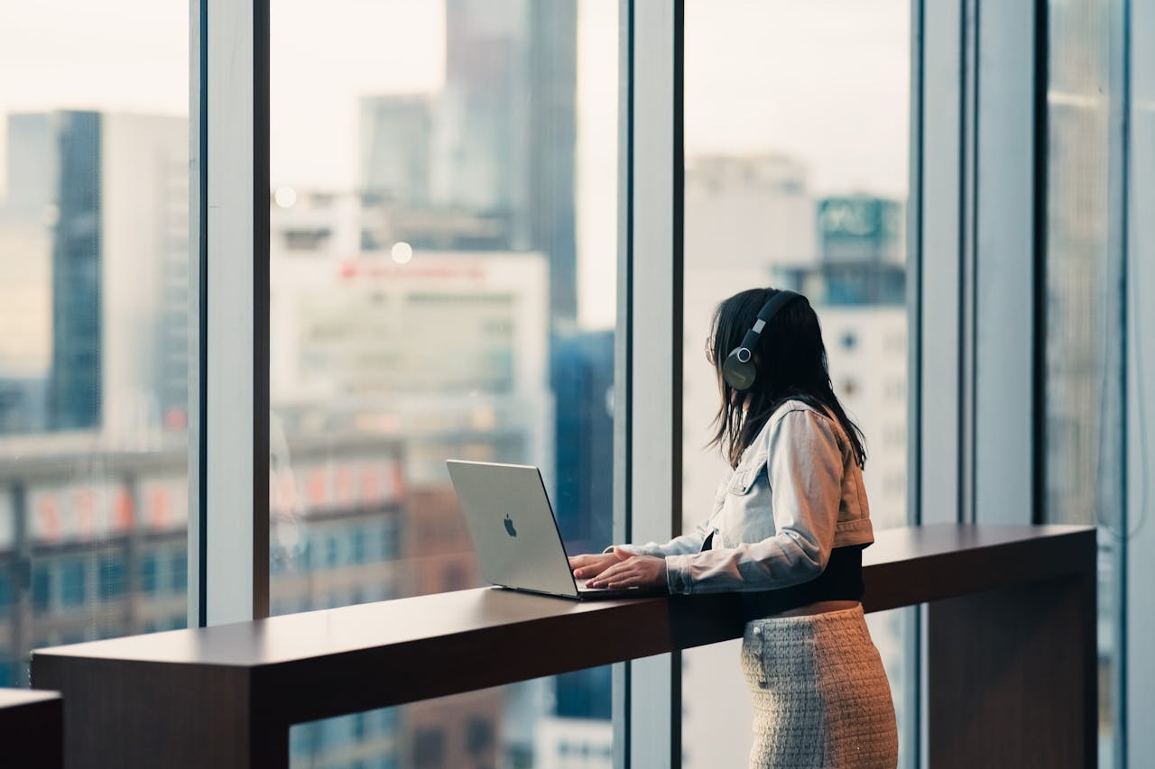 our-services-2 A woman wearing headphones is using a laptop in a modern office with city views.